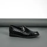 Mocasín de cuero vacuno negro con textura grabada tipo reptil. Presenta un diseño clásico con antifaz en el empeine, suela de goma en tono natural y taco bajo de madera.
