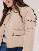 Campera corta capitoneada Michael Kors de color beige, con diseño acolchado en rombos, cuello camisero, cierre frontal de botones metálicos a presión y bolsillos frontales con solapa. Incluye un pequeño bolsillo en la manga.