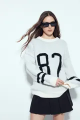 Sweater de punto color blanco con estampado numérico '87' en color negro en el frente y detalles de rayas horizontales en las mangas. Presenta cuello redondo y un corte relajado.