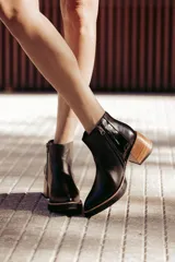 Botín de cuero negro, caña corta, con punta cuadrada y cierre lateral. Presenta un tacón de madera de altura media y detalles en charol con textura de cocodrilo en el talón.