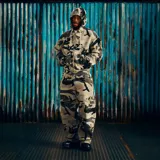 Campera con capucha de corte holgado, confeccionada en tela con estampado camuflado en tonos grises. Presenta un diseño gráfico bordado en la espalda que incluye un águila central, texto y detalles numéricos.