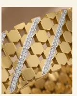 Pulsera tipo esclava rígida de acero con baño de oro, con diseño de malla calada y una línea central decorada con circonias brillantes.