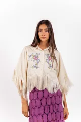 Camisa corta de color beige con silueta amplia, mangas anchas tipo capa terminadas en flecos y ramos de flores bordados en el frente.