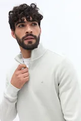 Buzo de algodón color beige claro, con cuello alto y cierre frontal parcial. Presenta tejido acanalado y un pequeño logo bordado en el pecho.