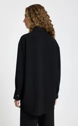 Camisa negra de tejido de punto con corte recto y ligeramente holgado. Presenta cuello estructurado, cierre frontal con botones, mangas largas con puños, bolsillo de parche delantero y bajo redondeado.