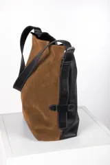 Bolso tipo bucket de diseño bicolor, confeccionado en una combinación de cuero gamuzado color tostado y cuero graneado negro. Presenta una correa ajustable con hebilla metálica estilo estribo y costuras a la vista.