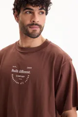Remera oversize de manga corta en color marrón, confeccionada en 100% algodón. Presenta un diseño gráfico estampado en la espalda con inscripciones en color blanco.