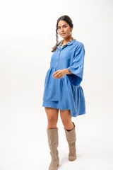 Vestido corto azul, estilo camisero, con cuello camisero, cierre frontal con botones y mangas acampanadas con volados. Presenta un diseño holgado con cortes fruncidos en el cuerpo.