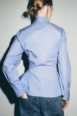 Camisa de algodón con cuello solapa, manga larga con puños y diseño peplum en el bajo. Presenta cierre frontal con botones ocultos por solapa.