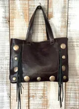 Bolso de hombro estilo boho, confeccionado en cuero marrón oscuro. Presenta un diseño rectangular con tachas metálicas circulares en los laterales y la base, además de flecos de cuero colgantes en ambos lados.