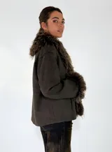 Chaqueta de textura gamuzada en color marrón, con solapas amplias y puños adornados con piel sintética de pelo largo en tono contrastante. Presenta un diseño de corte recto y estructura abierta.