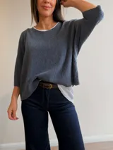 Conjunto de dos piezas compuesto por una remera básica blanca de algodón y un buzo liviano de textura plisada en color azul grisáceo. El buzo presenta un diseño de manga murciélago, corte holgado y detalles de hilo plateado en los bordes. Incluye un collar largo con dije circular de madera y metal.