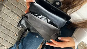 Cartera de hombro negra con acabado brillante, diseño rectangular con solapa frontal, cierre de hebilla metálica y detalles de tachas plateadas en los extremos inferiores.