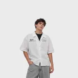 Camisa oversize blanca de manga corta, con cuello camisero, cierre frontal de botones y estampado tipográfico en el pecho.