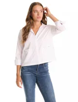 Camisa blanca de corte recto y estilo clásico, con cuello solapa, cierre frontal oculto y mangas largas con puños. Presenta dos bolsillos frontales decorados con apliques de tachas metálicas en el borde superior.