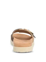 Sandalias tipo slide con tira ancha en tejido texturizado color beige y detalle de hebilla circular decorativa en tono marrón. Presentan una base ergonómica con suela de poliuretano ligera y flexible.