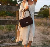 Cartera tipo bandolera de gamuza color marrón, con diseño rectangular y solapa frontal. Cuenta con un cierre metálico giratorio y correa larga ajustable para llevar al hombro o cruzada.