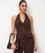 Chaleco de sastrería color marrón, con diseño de frente única y cuello halter. Presenta pinzas laterales que definen la silueta, bolsillos decorativos y ruedo levemente redondeado. En la espalda, cuenta con una cinta ajustable con ojal metálico y tiras largas.