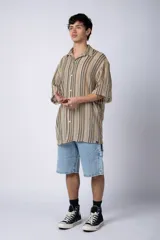 Camisa de manga corta con cuello solapa, corte holgado y cierre frontal de botones. Presenta un diseño de rayas verticales en tonos beige, negro y blanco.