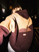 Campera con capucha de diseño patchwork, confeccionada en tejido tipo corderito en tonos marrón y tostado. Presenta un diseño que simula un paisaje montañoso con cortes irregulares y etiqueta de marca en la parte posterior.
