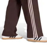 Pantalón deportivo Adidas de corte recto y estilo clásico, color marrón con franjas laterales en tono rosado. Cuenta con cintura elástica con cordón ajustable, bolsillos laterales con cierre y el logo del trifolio bordado en una de las piernas.