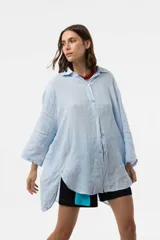 Camisa de lino color beige, de calce amplio y relajado, con cuello camisero, cierre frontal de botones, mangas largas y diseño de largo irregular con aberturas laterales.