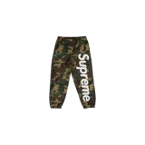 Pantalón de jogging Supreme de diseño camuflado en tonos verde militar, marrón y negro. Cuenta con elástico en la cintura y los tobillos, y lleva el nombre de la marca en grandes letras blancas aplicadas verticalmente en la pierna izquierda.