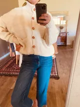 Cardigan tejido de lana color crudo, con cuello alto, cierre frontal con botones de madera, bolsillos delanteros y corte holgado.