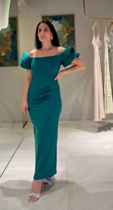 Vestido largo de fiesta color verde esmeralda, con escote recto estilo hombros caídos y mangas cortas con volados estructurados. Presenta un detalle de fruncido lateral en la cintura que aporta un ajuste entallado a la silueta.