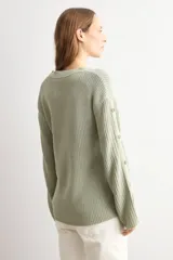 Sweater de punto color blanco natural, con cuello redondo y mangas largas adornadas con botones dorados a lo largo de las costuras exteriores.