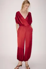 Mono largo satinado de color rojo, con escote en V cruzado y manga corta. Presenta cintura elástica con cinturón negro incorporado, bolsillos laterales ocultos y terminación de las piernas con puño elástico.