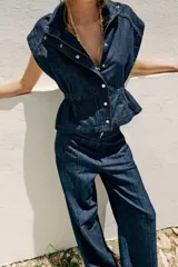 Chaleco de denim azul oscuro con cuello alto, manga sisa y cintura ajustable con elástico. Cierre frontal con botones a presión metálicos.
