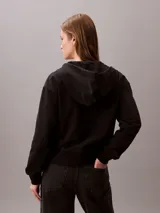 Buzo con capucha y cierre frontal de cremallera de la marca Calvin Klein, color negro, de corte regular cropped y manga larga. Presenta bolsillos tipo canguro, cordones ajustables en la capucha y el logo monograma de la marca estampado en el pecho.