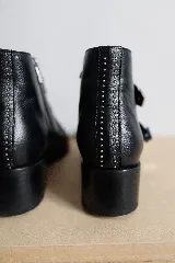 Botas cortas de cuero negro con puntera fina, taco bajo y cierre lateral. Presentan tres correas con hebillas plateadas y apliques de tachas plateadas.