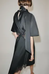Vestido midi de satén negro con cuello subido, manga corta tipo capa y pañuelo incorporado en el mismo tejido. Presenta aberturas laterales decoradas con encaje a tono y bajo asimétrico con detalles de encaje. Cierre en la espalda con abertura y botones forrados.