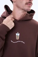 Buzo tipo canguro color marrón, de corte oversize, con capucha y bolsillo frontal tipo canguro. Presenta un pequeño estampado central con un vaso de café y una frase debajo.