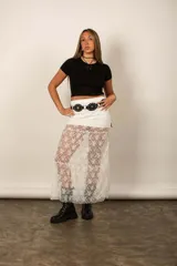 Pollera larga blanca de encaje transparente, con diseño de capas y cintura alta decorada con un cinturón negro de tachas.