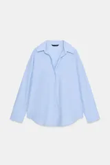 Camisa celeste de popelina, de corte holgado, con cuello solapa, escote en pico y mangas largas. Tiene bajo asimétrico y cierre frontal con botones.