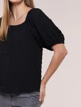 Blusa blanca de tela texturada y silueta fluida, con escote en U y frunces. Cuenta con mangas cortas abullonadas con elástico en los puños y ruedo redondeado.