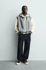 Cazadora estilo varsity o bomber, cuerpo principal gris oscuro y mangas color crema. Presenta cuello, puños y bajo en rib elástico con rayas grises y blancas. Tiene bordados en el pecho: un monograma estilizado a la izquierda y el texto "EST. 1975" a la derecha.