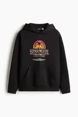 Canguro negro con capucha y bolsillo frontal tipo canguro. Presenta un estampado gráfico en el pecho con el logo de la serie Stranger Things y una silueta del Mind Flayer sobre un atardecer.