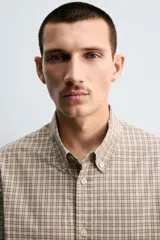 Camisa de algodón de manga larga, cuello abotonado y cierre frontal de botonadura. Presenta un patrón de cuadros pequeños en tonos beige, marrón y blanco.