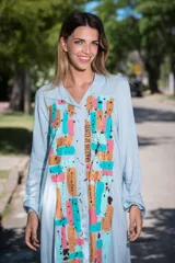 Vestido camisola largo de color celeste pastel, confeccionado en viscosa, con mangas largas y cuello camisero. Presenta una estampa frontal pintada a mano con diseño abstracto de pinceladas en tonos dorado, cobre, turquesa y nude, incluyendo la firma de la artista Carolina De Cunto.