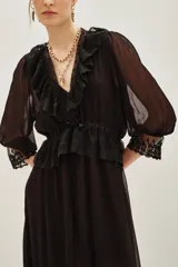 Vestido midi negro de estilo romántico, confeccionado en tejido semitransparente con detalles de encaje en el cuello, la cintura y el ruedo. Presenta mangas tres cuartos con terminación en encaje, escote en V con volados y un lazo de ajuste en la cintura.