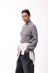 Polera de tejido de punto color marrón, con cuello alto y diseño de trenzas verticales. Presenta mangas largas con puños ajustados y un detalle de panel lateral asimétrico que se anuda en la cintura.