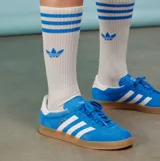 Championes Adidas Gazelle Indoor de gamuza azul con las icónicas tres rayas laterales en blanco. Presenta una suela de caucho color caramelo, talón blanco y detalles dorados con el nombre del modelo en el lateral.