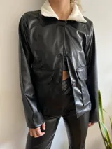 Chaqueta negra de cuero sintético con cuello de corderito blanco, corte recto, cierre con botones y bolsillos frontales con solapa.