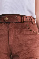 Short corto de tiro bajo confeccionado en gamuza elastizada color marrón camel. Presenta detalles de tachas metálicas doradas en los bolsillos delanteros y en los bordes inferiores, además de una faja en la cintura con hebillas metálicas laterales.
