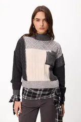 Sweater de punto con diseño patchwork en tonos gris, grafito y crudo, con cuello alto tejido. Presenta una estructura de bloques de color y texturas variadas, incluyendo detalles de trenzas y tejido acanalado.