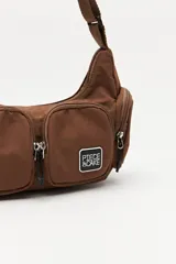 Cartera tipo shoulder color beige, de diseño utilitario y formato curvo. Confeccionada en material liviano y resistente, cuenta con correa regulable para llevar al hombro o cruzada. Presenta múltiples bolsillos con cierre en el frente y laterales, y un parche con el logo "Piece & Cake" en el frente.